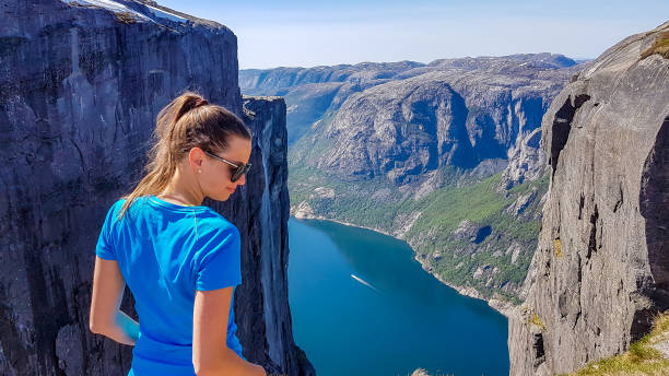 girl wearing blue t-shirt looks down, past her shoulder. she is enjoying the fjord view in front of her. stunning lysefjorden shimmers with many shades of blue and green. girl is happy after the hike - kjeragbolten imagens e fotografias de stock