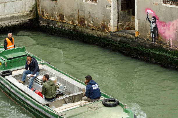 VENICE, ITALY - MAY 15, 2019:  Four men on a contruction workers' boat travelling past the stencil street art depiction of a child wearing a lifejacket and waving a flare attributed to graffiti artist Banksy.  Viewed from a public street overlooking the Rio di Ca'Foscari in the Dorsoduro student district of Venice.