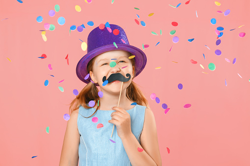 Happy father's day. Funny little girl with fake mustache and wearing a hat under falling confetti on a pink background. Family concept