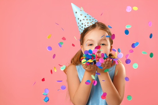 Cheerful little girl celebrates birthday. The child blows confetti from the hands. Closeup portrait on pink coral background.