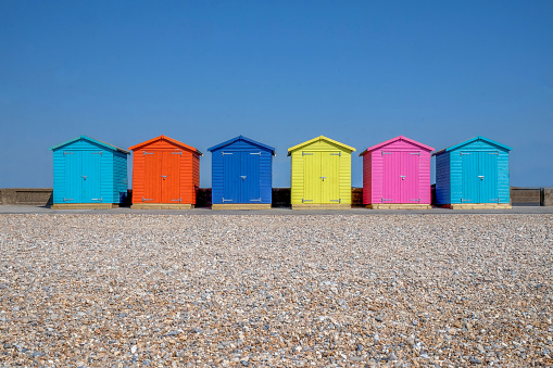 A line of six brightly coloured Beach huts, each one is a different colour in the foreground is a pebble beach and behind is a bright blue sky, Seaford, east Sussex, England, United Kingdom, UK,