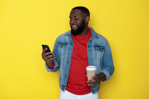 Half length of happy American guy in denim jacket standing against yellow background. He is looking at screen of mobile phone and drinking coffee in studio