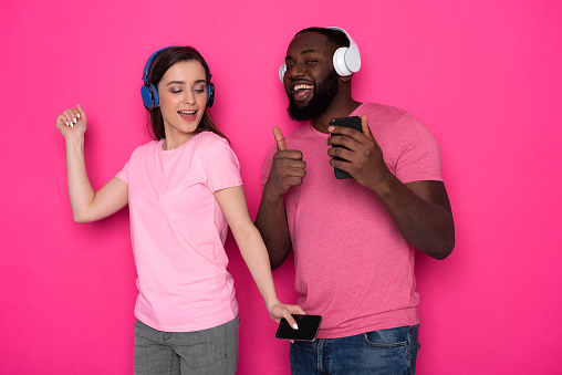 Half length of happy African man dancing with Caucasian pretty lady and showing thumb up against pink background. Smiling American guy with his girlfriend listening to music and holding smartphones in arms. Modern technologies concept