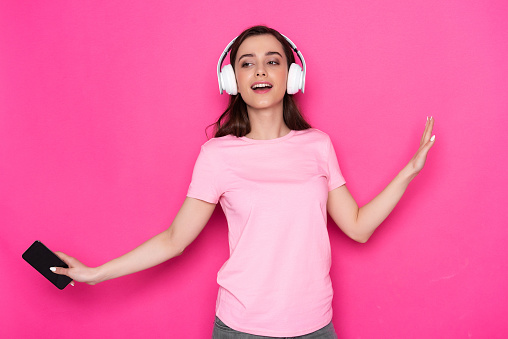 Waist up of happy young lady wearing casual and holding mobile phone in arm against pink background. She is enjoying her favourite music in white earbuds while gesticulating with hands in studio