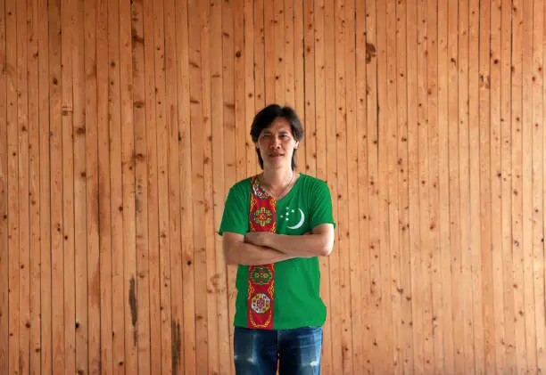 Photo of Man wearing Turkmenistan flag color shirt and cross one's arm on wooden wall background.