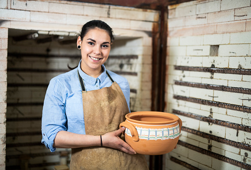 Portrait of smiling young woman in pottery studio, Portrait of young Latino woman in ceramic studio holding clay vessel.