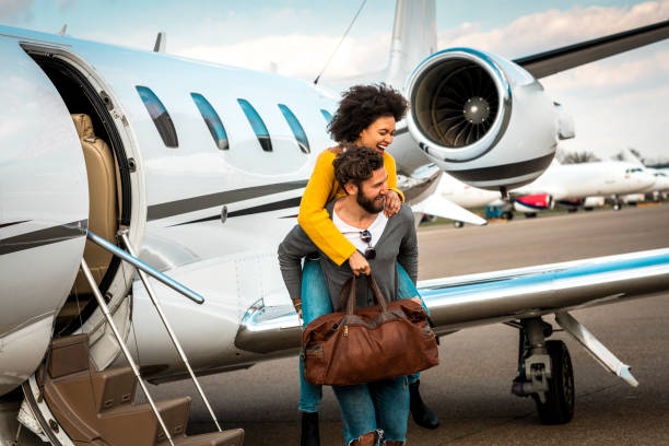 joven exitosa mujer abrazando amorosamente a su pareja desde atrás junto a un avión privado estacionado en una pista de aterrizaje del aeropuerto - status symbol audio fotografías e imágenes de stock