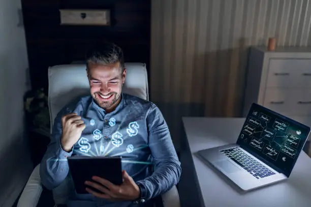 Photo of Happy young handsome businessman holding tablet and cheering for earning lots of money. On laptop monitor is business plan.