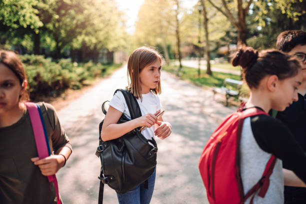 Confused schoolgirl standing in schoolyard with classmates Confused schoolgirl wearing backpack standing alone in schoolyard beside a group of classmates and holding smartphone school exclusion stock pictures, royalty-free photos & images