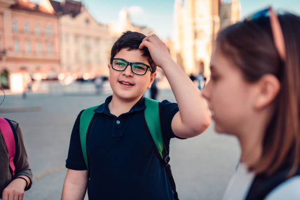 School boy wearing eyeglasses and backpack hang out with friends on town square and scratching head