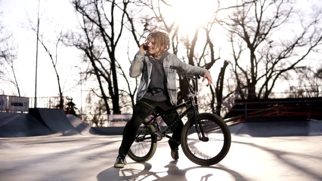 Portrait of happy handsome teenage boy with dreadlocks, using mobile phone while riding a BMX bike, smiling, messaging friends via social networks. People, lifestyle and communication concept. Low angle footage
