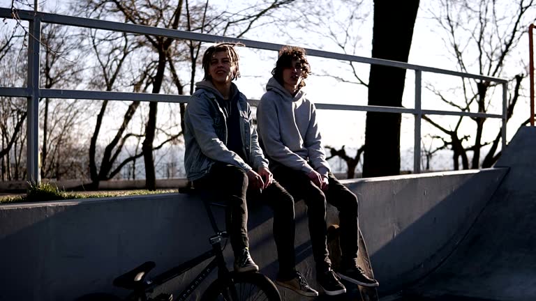 Two teenage men are sitting on high parapet together. Bmx rider and skateboarder - a boy with the dreadlocks is openly smoking. Bad habit. Street life