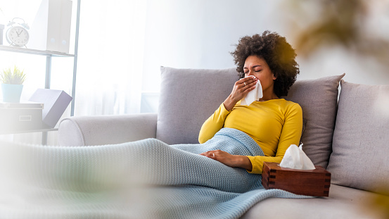 Sick young woman sitting on sofa blowing her nose at home in the sitting room. Photo of sneezing woman in paper tissue. Picture showing woman sneezing on tissue on couch in the living-room