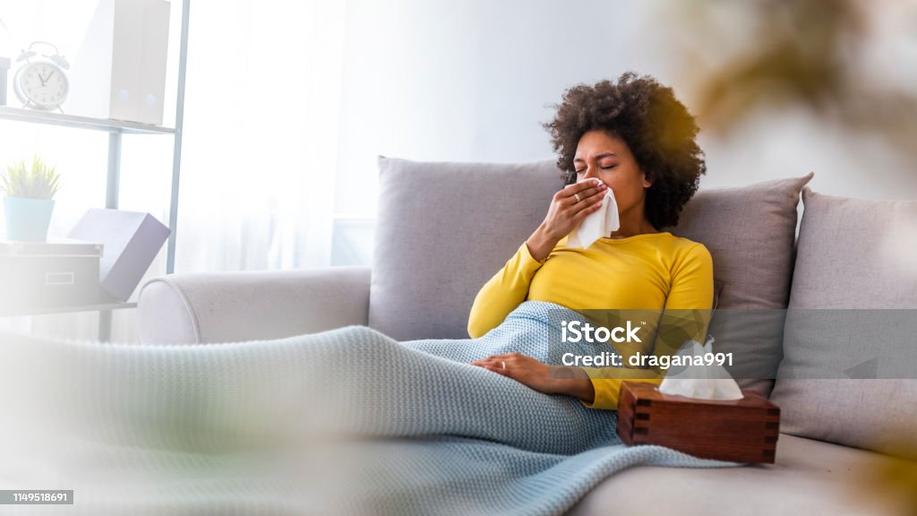 Woman sneezing in a tissue in the living room. Sick young woman sitting on sofa blowing her nose at home in the sitting room. Photo of sneezing woman in paper tissue. Picture showing woman sneezing on tissue on couch in the living-room Cold And Flu Stock Photo