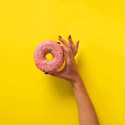 Female hand holding pink donut over yellow background. Top view, flat lay. Sweet, dessert, diet concept. Banner with copy space. Weight lost after holidays.