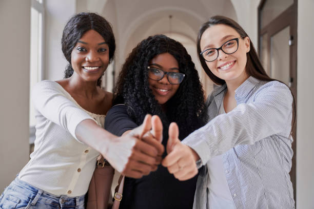 tre studentesse di razza mista felici sorridenti, guardando la macchina fotografica e mostrando il cartello super per pollice in su nel luminoso corridoio universitario. concetto di importanza dell'educazione e dello studio nella nostra vita - skirt women jeans white foto e immagini stock