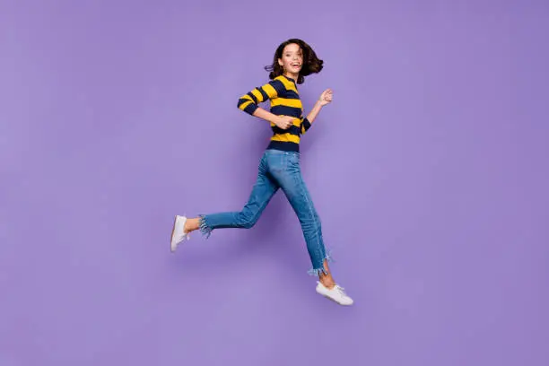 Photo of Full length side profile body size photo beautiful she her lady cheerful jump high flight hurry shopping sale discount mall store wear blue yellow striped pullover isolated violet purple background