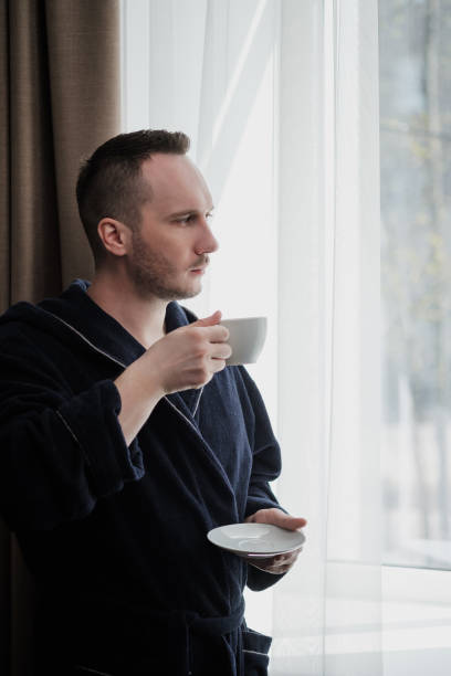 istock Young brunette man in a blue robe by the window. Drinks coffee or tea from a white cup with a saucer. Morning, sunshine, bedroom window. 1149530231