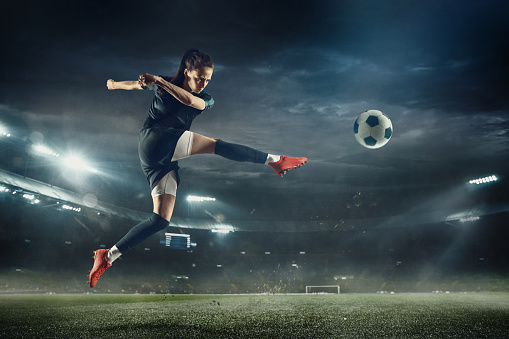 Young female soccer or football player with long hair in sportwear and boots kicking ball for the goal in jump at the stadium. Concept of healthy lifestyle, professional sport, hobby, motion, movement.