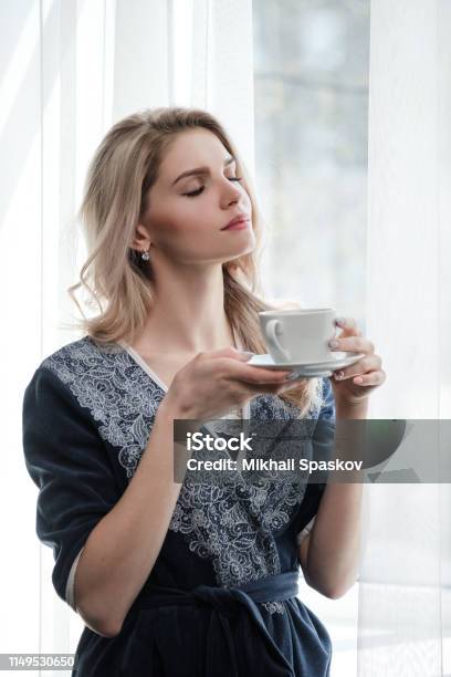 Beautiful Young Blonde Woman In A Blue Robe By The Window Drinks Coffee Or Tea From A White Cup With A Saucer Morning Sunshine Bedroom Window Stock Photo - Download Image Now