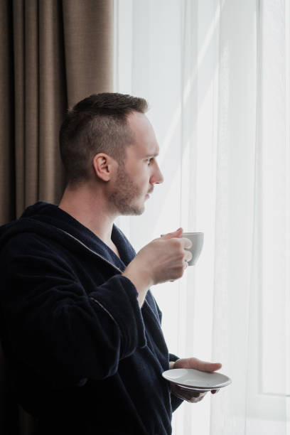 istock Young brunette man in a blue robe by the window. Drinks coffee or tea from a white cup with a saucer. Morning, sunshine, bedroom window. 1149531077