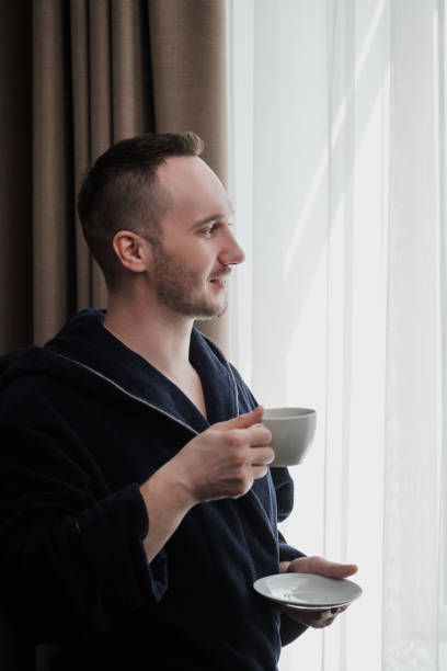 istock Young brunette man in a blue robe by the window. Drinks coffee or tea from a white cup with a saucer. Morning, sunshine, bedroom window. 1149531316