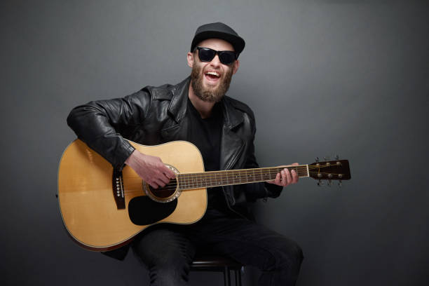 istock Guitar player singing in music studio. Hipster guitar player with beard and black clothes playing the acoustic guitar 1149534285