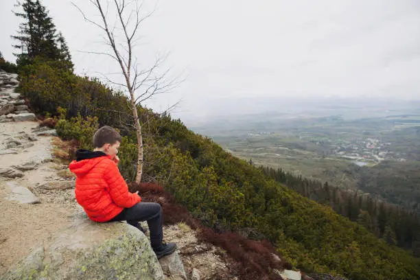 Stary Smokoves, Slovakia - April 28, 2019: teenager boy is taking a break and having lunch while sitting on a big rock in beautiful mountains.