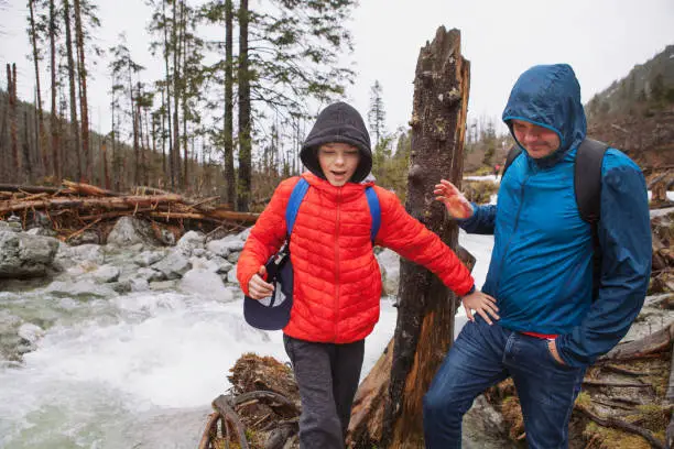 Stary Smokoves, Slovakia - April 28, 2019: Two backpackers - father and teenager son having fun in the rain in the mountains next to a river and tree trunk