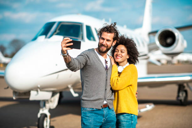 pareja multiétnica tomando un selfie en un teléfono móvil frente a un jet privado estacionado en una calle de rodaje del aeropuerto - status symbol audio fotografías e imágenes de stock