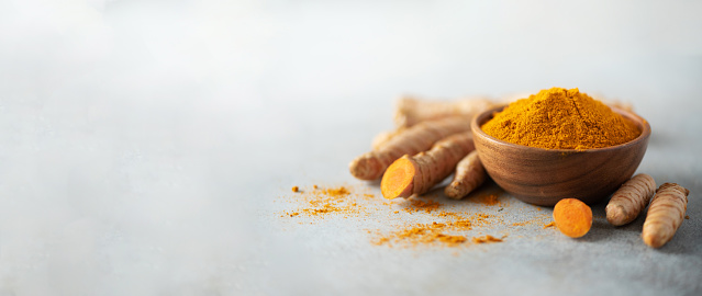 Turmeric powder in wooden bowl and fresh turmeric root on grey concrete background. Banner with copy space.