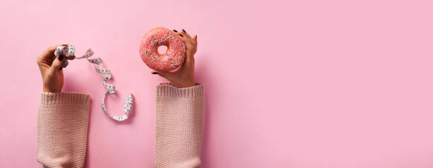 les mains femelles retenant le donut et le ruban de mesure sur le fond rose. vue de dessus, plat lay. doux, dessert, concept de régime. bannière avec espace de copie. poids perdu après les vacances - weight women medicine lost photos et images de collection