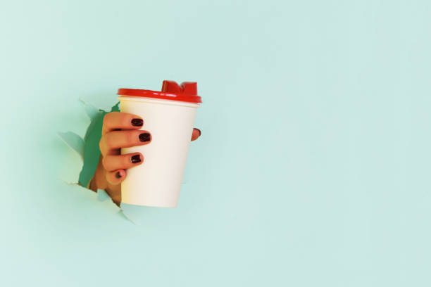 Female hand holding white paper mug on blue background. Take away coffee cup concept. Mock up with copy space