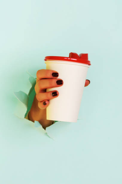 Female hand holding white paper mug on blue background. Take away coffee cup concept. Mock up with copy space
