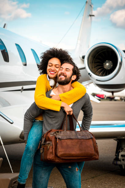 jeune femme célèbre amoureusement étreignant son partenaire de derrière le prochain jet privé stationné sur une voie de circulation d’aéroport - status symbol audio photos et images de collection