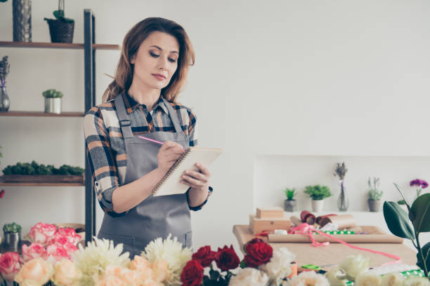 istock Portrait focused concentrated lady vendor use make note pen diary organizer minded pensive thoughtful flora assortment wavy curly hairdo casual style trendy stylish checkered shirt outfit indoors 1149570177