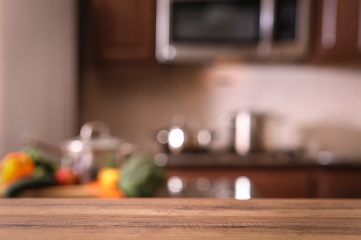 Defocused modern kitchen with empty wooden table or countertop in foreground for object placement.  Cooking pans and food preparation in background.  Stainless steel appliances. No people.