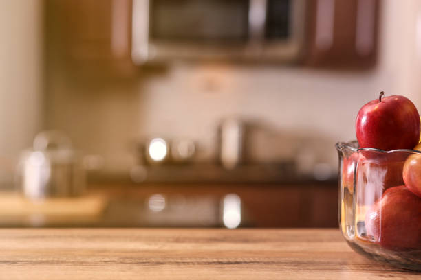 Defocused modern kitchen with apples in bowl on wooden table or countertop in foreground.  Cooking pans in background.  Stainless steel appliances.  No people.