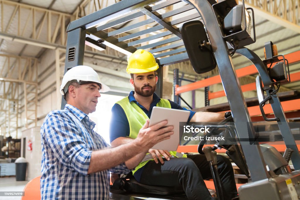 Manager showing digital tablet to manual worker Mature manager showing digital tablet to colleague in industry. Production worker is sitting on forklift. They are working together. Forklift Stock Photo