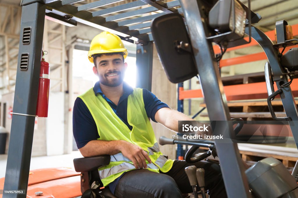 Smiling male engineer on forklift in factory Smiling male engineer working in factory. Skilled production worker sitting on forklift. He is in distribution warehouse. Forklift Stock Photo