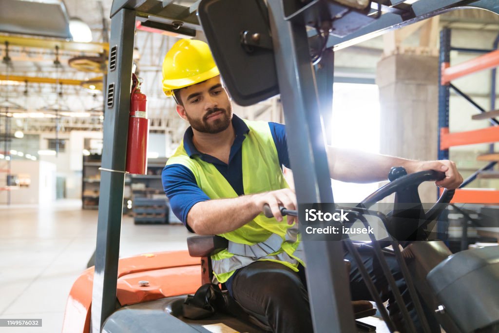 Bearded young man driving forklift in factory Bearded young man driving forklift in factory. Manual worker is working in warehouse. He is occupied in manufacturing. Forklift Stock Photo