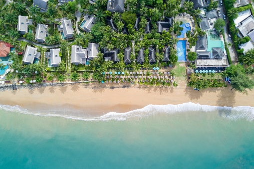 Aerial view of resort villa layout with coconut trees, umbrellas and deck chairs on the beach. Ocean wave reaching coastline. Beautiful tropical beach from top view. Andaman sea in Thailand. Summer holiday concept