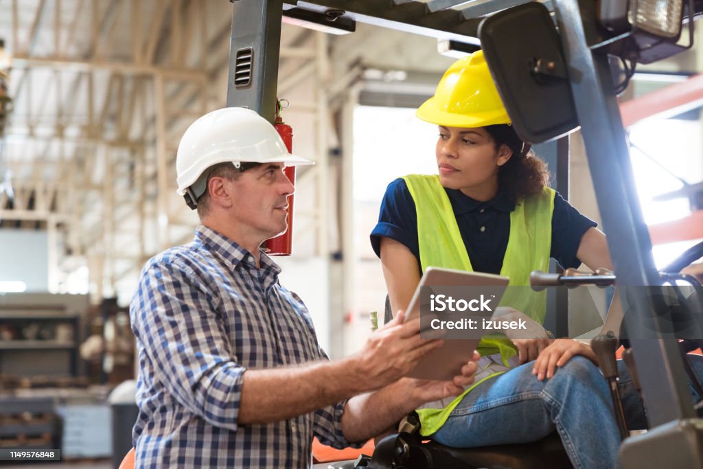 Forklift driver looking at manager in industry Manager and female engineer discussing over digital tablet. Forklift driver is looking at foreman. They are in manufacturing industry. Manufacturing Occupation Stock Photo
