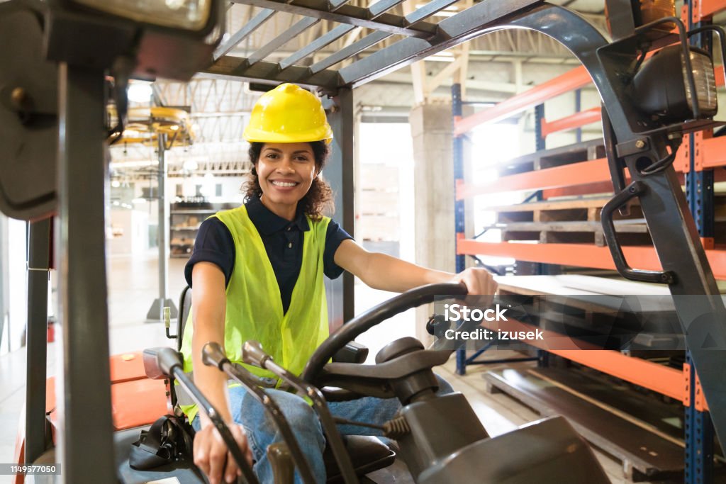 Smiling female worker sitting on forklift Portrait of smiling female worker sitting on forklift. Confident mid adult engineer is working in manufacturing factory. She is wearing hardhat and reflective clothing. Forklift Stock Photo