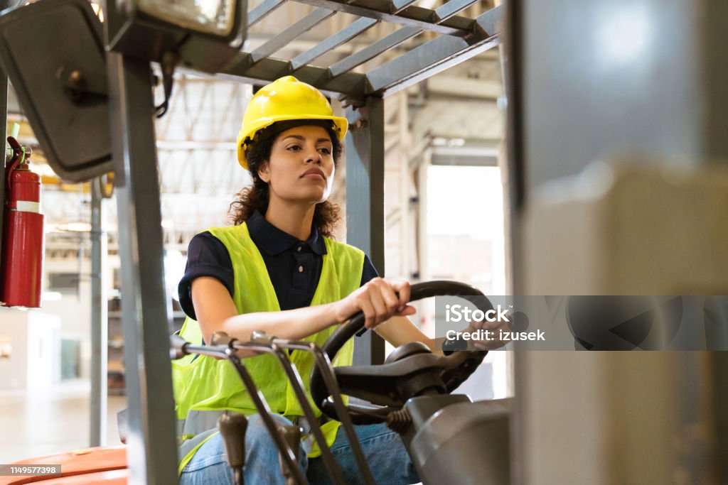 Confident female driving forklift in factory Confident female driver driving forklift. Mid adult woman is working in distribution warehouse. She is wearing hardhat and reflective clothing in factory. Forklift Stock Photo
