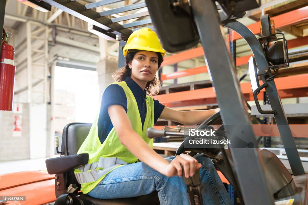Female worker driving forklift in industry Mid adult female worker driving forklift in industry. Engineer is occupied in manufacturing sector. She is with confident look on her face. Driver - Occupation Stock Photo