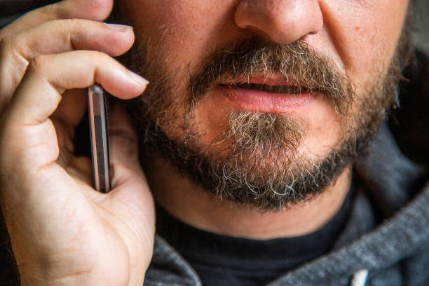 Portrait of man threating someone over the phone Close up portrait of bearded man talking on smart phone, focus on lips, no eyes, secret talk, criminal threating someone hoax stock pictures, royalty-free photos & images