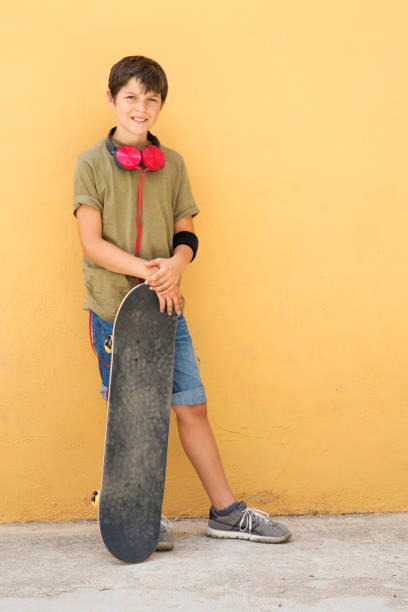 istock Young boy leaning on a yellow wall with headphones on neck, holding a skateboard while looking away in a bright day 1149603374