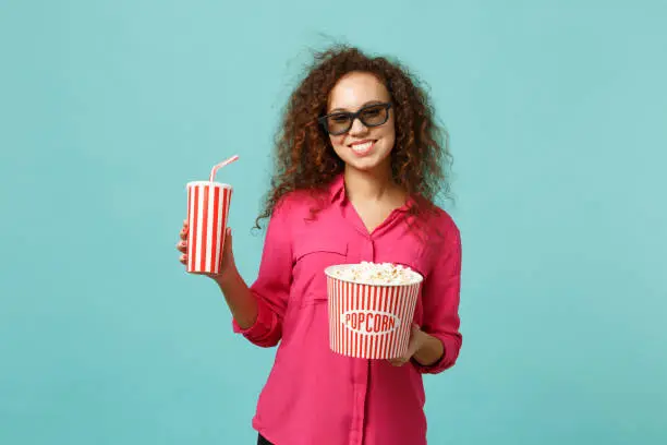 Smiling african girl in 3d imax glasses watching movie film hold popcorn, cup of soda isolated on blue turquoise background in studio. People emotions in cinema, lifestyle concept. Mock up copy space