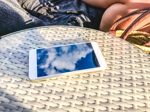 Reflection of blue sky with clouds in the phone-smartphone, which lies on a bright wicker table against the background. Blur and abstraction